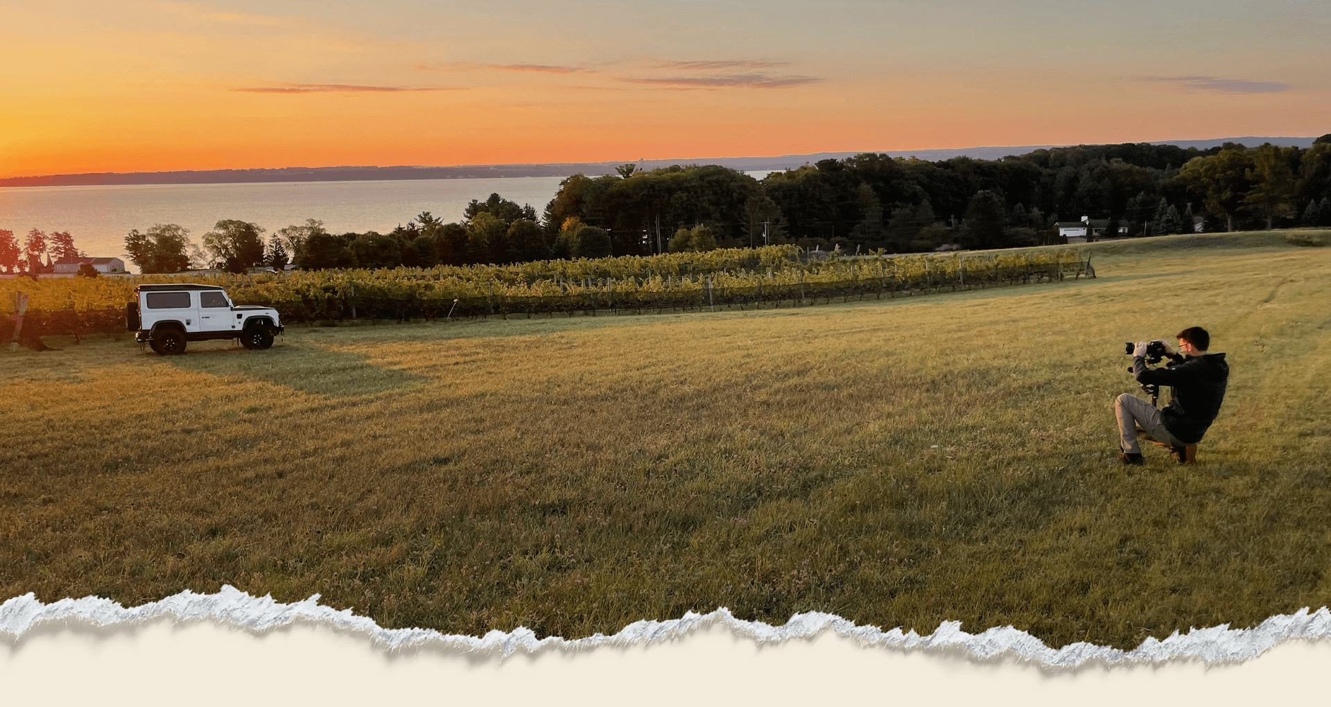 Photographer filming a Land Rover beside a vineyard at sunset over the bay